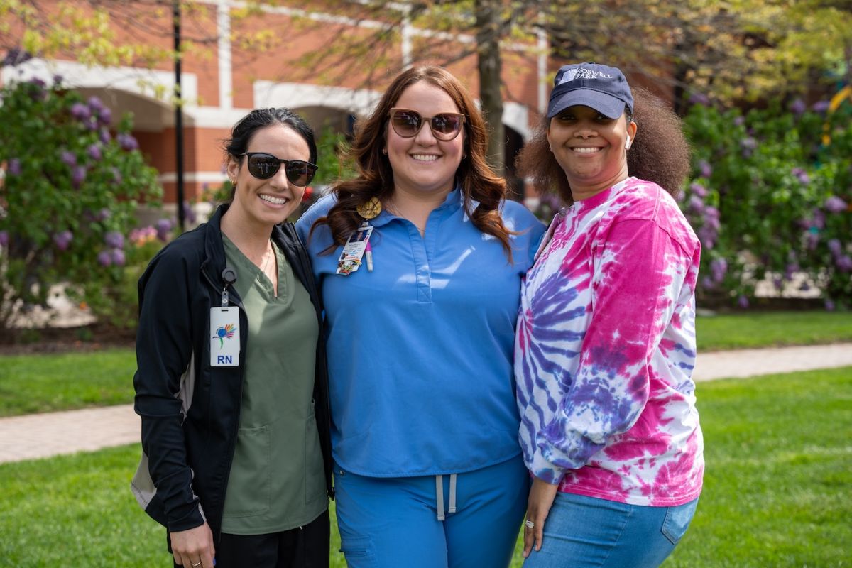 Three women smiling as they stand in a group together. Two are in medical scrubs and one has a tie-dye T-shirt on.