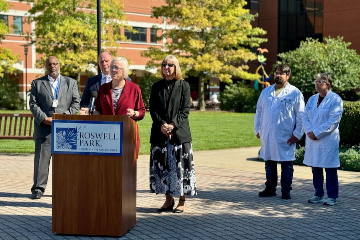 Woman in red stands at podium in outside setting with people standing behind her