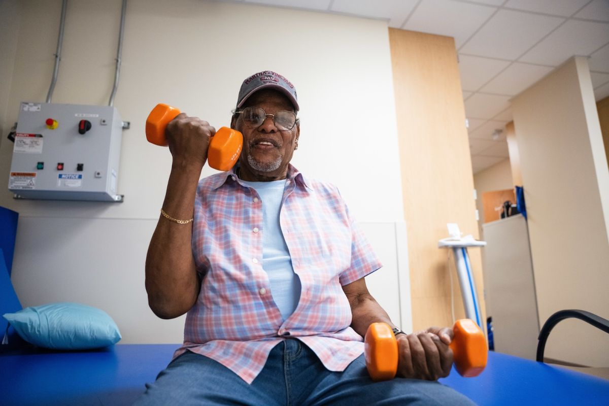 A patient sits on a blue bench lifting orange hand weights as part of his physical therapy. 