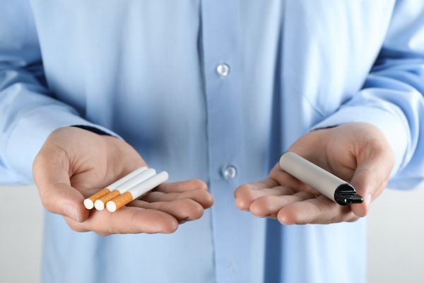 Stock image showing midsection of person in blue dress shirt holding cigarettes in one hand and a vape in the other